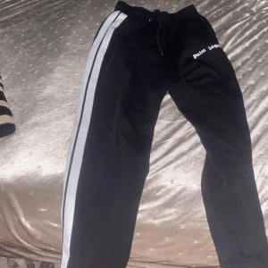 Palm Angels svarta mjukisbyxor - Svarta mjukisbyxor från Palm Angels med vita sidoremsor och logotyp på vänster höft. Byxorna har snörning i midjan och en avslappnad passform, perfekta för en chill och sportig look.