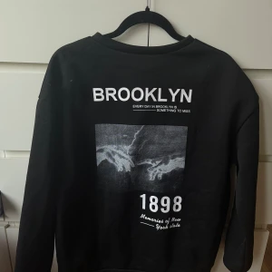 Svart sweatshirt med Brooklyn-tryck - Svart sweatshirt från Shein med stort vitt Brooklyn-tryck på ryggen, coolt grafiskt motiv och texten '1898 Memories of New York state'. Tröjan har rund hals och långa ärmar, perfekt för en streetstyle-look.