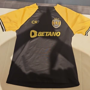 Sporting CP CR7 Nike fotbollströja M - Sporting CP fotbollströja i svart och gult med CR7-tryck, klubbmärke och sponsorloggor. Tillverkad i Nike Dri-FIT-material som andas. Kortärmad modell med snygga kontrastärmar och broderad klubbsköld på bröstet. 