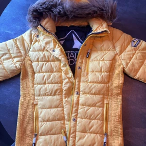 Gul pufferjacka med fuskpäls från Sublevel - Snygg gul pufferjacka från Sublevel med fuskpäls på huvan och quiltad design. Jackan har dragkedjor i kontrastfärg, flera fickor och en badge på ärmen. Perfekt för kalla dagar och ger en cool streetstyle-vibe.