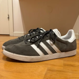 Adidas Gazelle grå/vita sneakers - Säljer ett par klassiska Adidas Gazelle sneakers i grå mocka med vita detaljer och svarta snören. Skorna har de ikoniska tre ränderna på sidan, vit hälkappa med svart Adidas-logo och platt sula. Perfekta för dig som gillar retrostil och streetwear. Knappt använda.