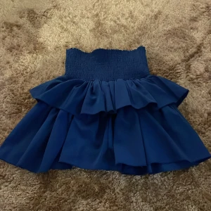 Mörkblå volangkjol med smock - Supersöt mörkblå kortkjol med bred smock i midjan och dubbla volanger. Kjolen har en flowy känsla och passar perfekt till en trendig look. Materialet känns lätt och luftigt, och den höga midjan ger en snygg siluett.