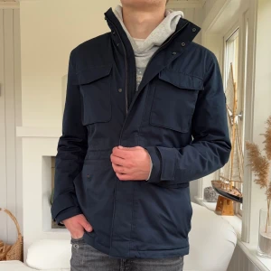 Mörkblå fieldjacket  från J.Lindeberg - Säljer en stilren mörkblå fieldjacket från J.Lindeberg i storlek M. Jackan har fyra stora fickor framtill, hög krage och dragkedja med tryckknappar. Perfekt för dig som vill ha en clean look med funktionella detaljer.