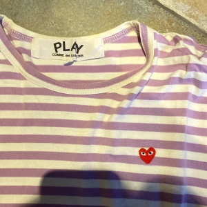 långärmad tröja från Comme des Garçons - Långärmad tröja från Comme des Garçons Play med lila och vita ränder. Klassisk rund halsringning och ikoniskt rött hjärta broderat på bröstet. Skön och lätt i bomull, perfekt för dig som gillar stilrena men lekfulla plagg.