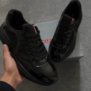 Svarta sneakers - Säljer ett par svarta  sneakers med glansiga detaljer och meshpaneler. Skorna har rund tå, snörning och en diskret röd logga på plösen. Snygg och modern design som sticker ut med sin kombination av material och helsvart look.