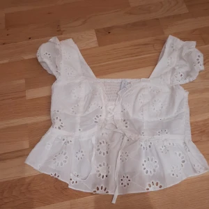 Vit blus med broderade blommor Amisu - Superfin vit blus från Amisu med broderade blommor och volangdetaljer. Toppen har korta puffärmar, snörning framtill och är tillverkad i bomull. Perfekt för sommarens alla tillfällen!