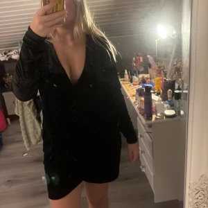 Svart sammetsklänning med v-ringning - Säljer en svart sammetsklänning med djup v-ringning och lång ärm. Klänningen är kort och har en omlottkänsla framtill som ger en snygg siluett. Perfekt för dig som vill sticka ut med en elegant och trendig look.