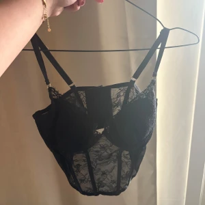 Svart spetskorsett med justerbara band - Säljer en svart korsett med genomskinlig spets och justerbara axelband. Toppen har snygga detaljer med mesh och spets, samt markerade kupor för extra stöd. Perfekt för dig som vill ha en edgy och trendig look.