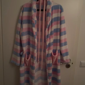 Badrock  - Mysig och lång badrock i fleece med breda ränder i ljusrosa, ljusblå och vitt. Badrocken har två fickor, knytband i midjan och en skön huva. Perfekt för att chilla hemma eller hålla sig varm under kyliga dagar.