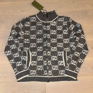 Grå kofta Gucci kofta - Exklusiv grå kofta från Gucci med vit GG-logga över hela tröjan. Koftan har ribbade muddar, ståkrage och hel dragkedja framtill. Tillverkad i 100% ull för en lyxig och mjuk känsla. Snygga vita detaljer vid krage, ärmslut och nederkant.