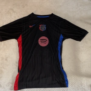 FC Barcelona Lamine Yamal svart matchtröja - Säljer en svart FC Barcelona fotbollströja från Nike med Lamine Yamal och nummer 19 på ryggen. Tröjan har röda och blå detaljer på sidorna, klubbmärke och Spotify-logga på bröstet. Tillverkad i lätt och ventilerande material, perfekt för match eller träning.