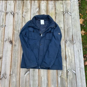 J.Lindeberg field jacket  - Snygg marinblå field jacket från J.Lindeberg med dragkedja och knappar framtill, två stora bröstfickor och hög krage. Jackan har en stilren look med diskret logotyp på ärmen och är tillverkad i lätt polyester. Perfekt för höst och vintern.