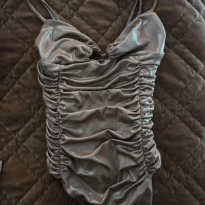 Glittrig guld body med rynk - Supersnygg glittrig guldfärgad body med smala axelband och rynkade detaljer längs sidorna. Perfekt för dig som vill ha något extra till sommaren!