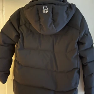 Svart pufferjacka Sail Racing 160 - Svart pufferjacka från Sail Racing med Sail Antarctica Expedition-märke på ärmen. Jackan har huva, breda kanaler och hög krage. Perfekt för kalla dagar och har en clean, sportig look. Materialet är syntet och jackan är långärmad.