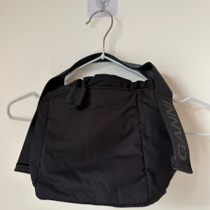 Svart axelväska från Ganni - Svart axelväska från Ganni med bred axelrem där loggan är tryckt. Väskan har en mjuk, puffig form och är tillverkad i 100% återvunnen polyester. Perfekt storlek för det viktigaste och har en enkel, stilren look.