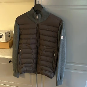 Moncler Jacka - Snygg jacka från Moncler i mörkgrått med quiltad front och stickade ärmar och rygg. Jackan har hög krage, två dragkedjefickor och Moncler-logga på ärmen. Perfekt för kyliga dagar när du vill hålla stilen.
