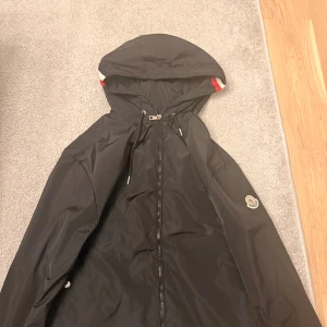 Svart vindjacka från Moncler med huva - Svart vindjacka från Moncler med huva och dragsko. Jackan har dragkedja framtill och Moncler-logga på ärmen. Huvan har en snygg detalj med röd, vit och blå rand. Perfekt för dig som vill ha en stilren och sportig look.