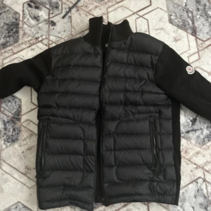 Svart Moncler pufferjacka med stickade ärmar - Svart pufferjacka från Moncler med quiltad front och rygg i glansigt material samt stickade ärmar. Jackan har dragkedja framtill, två fickor med dragkedja och Moncler-logga på ena ärmen. Perfekt för dig som vill ha en stilren och sportig look.