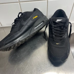 Nike Air Max 90 svart/gul sneakers - Svarta Nike Air Max 90 sneakers med gula detaljer och klassisk Air Max-dämpning. Skorna har en mix av mesh och skinn, snörning och en robust platt sula. Swoosh-logga på sidan och Air Max-tryck i gult vid hälen. Perfekta för dig som gillar streetwear och sportig stil.