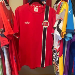 Röd Norge fotbollströja 2012/13 från Umbro Äkta - Säljer en röd Norge fotbollströja från Umbro i storlek L. Tröjan har korta ärmar, rund hals och ett tryckt Umbro-logo på bröstet. På framsidan finns Norges flagga och en svart-vit rand som går vertikalt. Materialet är lätt och andas, perfekt för match eller träning.  Äkta