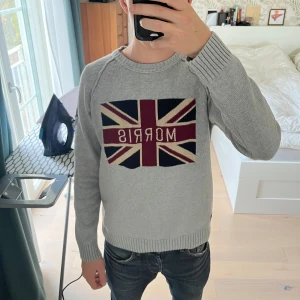 Grå stickad Morris tröja med flagga - Snygg grå stickad tröja från Morris med Union Jack-flagga och broderad logga på bröstet. På ena ärmen finns svart broderi med texten 'MG 1968'. Tröjan har rund hals, ribbade muddar och lång ärm. Perfekt för dig som gillar klassisk stil med en twist. Storlek M men passar S, jag är 177cm och väger 62kg, passar väldigt bra på mig