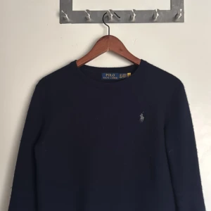 Mörkblå stickad tröja Polo Ralph Lauren - Mörkblå stickad tröja från Polo Ralph Lauren med klassisk rund hals och broderad logga på bröstet. Tröjan har lång ärm och är tillverkad i mjukt material som passar perfekt för kyligare dagar.