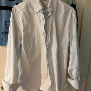 Randig skjorta från These Glory Days - Snygg skjorta med smal passform från These Glory Days. Skjortan är vit med ljusblåa tunna ränder, har klassisk krage och knappar framtill. Tillverkad i mjuk bomull och har långa ärmar. Perfekt för dig som gillar stilrena och fräscha plagg. (OANVÄND)
