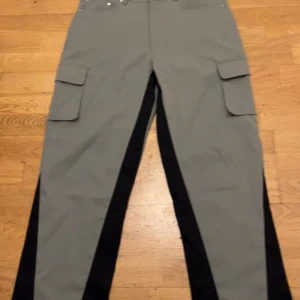 Grå cargopants från BoohooMAN - Snygga grå cargopants från BoohooMAN med svarta paneler längs benen. Byxorna har stora fickor på sidorna och klassisk midja med knappstängning. Perfekt för dig som gillar streetwear och vill ha en cool look.
