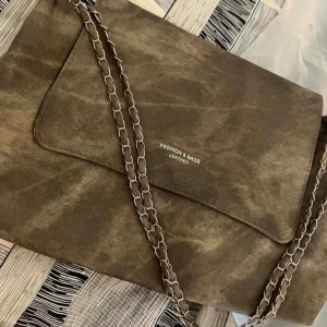 Brun axelväska med kedjerem - Stilren classy brun axelväska med kedjerem i silverfärg. Väskan har texten ”FASHION & BAGS LEATHER”  (obs den är ej i läder). Väskan har ett mjukt, mockaliknande material och enkel design med lock som är magnetisk. Rymlig insida som rymmer allt du behöver till vardagen. Perfekt för att exempelvis ha skolböcker i. (Fler bilder kan skickas om de behövs) Väskan har använts och kan ha små tecken på användning men inget direkt märkbart.