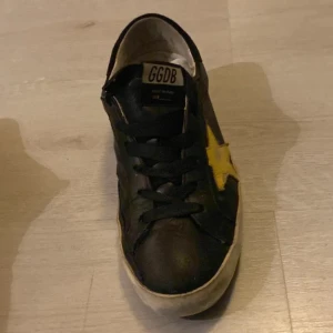 Svarta GGDB sneakers med gul stjärna - Säljer ett par svarta sneakers från Golden Goose med en stor gul stjärna på sidan. Skorna har snörning, vit platt sula och GGDB-logga på plösen. Tillverkade i skinn och har en rund tå. Perfekt för dig som gillar streetstyle och coola detaljer.