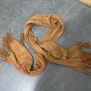 Brun halsduk med blommor och paljetter - Snygg brun halsduk med fransar i ändarna och broderade blommor dekorerade med guldiga paljetter. Mjuk och skön, perfekt att slänga över axlarna för en extra touch. Blommönstret och detaljerna gör den unik och trendig.