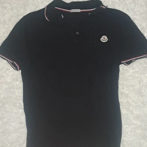 Svart Moncler pikétröja med logga - Svart pikétröja från Moncler med klassisk krage och knappar framtill. Tröjan har Moncler-logga på bröstet och rödvit randig detalj på krage och ärmslut. Perfekt för en stilren och sportig look.