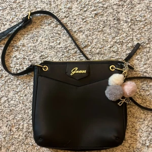 Svart axelväska från Guess med pompom - Säljer nu en äkta Guess väska som jag fixk av min mamma i present för några år sedan, därav inget kvitto. Snygg svart axelväska från Guess i skinnimitation med guldfärgade detaljer och dragkedja. Väskan har en kedja med fluffiga pompoms i beige, grått och ljusrosa samt ett glittrigt Guess-hänge. Insidan är fodrad med mönstrat tyg och har flera fack.