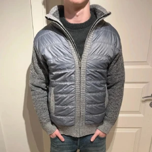 Grå quiltad cardigan jacka - Säljer en grå cardigan jacka med quiltad front i syntetmaterial och stickade ärmar samt sidor. Jackan har dragkedja och hög krage, perfekt för kyliga dagar. Snygg kombination av stickat och glansigt tyg som ger en modern look.