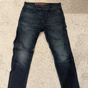 Mörkblå tapered jeans - Snygga mörkblå jeans med tapered fit och klassiska orange sömmar. Jeansen har fem fickor, subtila slitningar och en stilren look. Perfekta för dig som gillar en smalare passform utan att de sitter för tight. Materialet är klassisk jeansbomull.