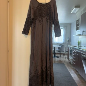 Brun långklänning med broderi - Säljer en brun långklänning med vackra broderade detaljer på ärmar, bröst och nederkant. Klänningen har trekvartsärmar och ett bohemiskt mönster som ger en unik look. Perfekt för dig som gillar avslappnad och stilren design.