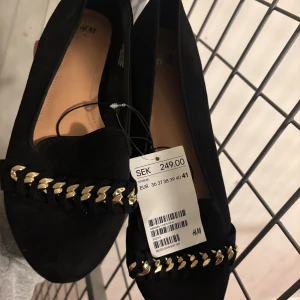 Svarta loafers med guldkedja från H&M - Snygga svarta loafers från H&M med platt sula och rund tå. Skorna har en dekorativ guldfärgad kedjedetalj framtill som ger en cool touch. Tillverkade i mockaimitation för en mjuk och stilren känsla. Perfekta att lyfta din outfit med.