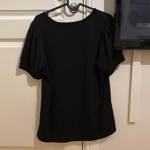 Svart blus med puffärm och knappar - Svart blus med puffiga korta ärmar och knappar vid ärmslutet. Ring halsringning och är gjord i ett mjukt, slätt material. St L (sitter som M)