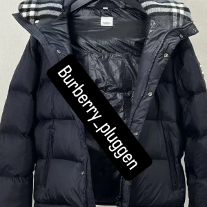 Burberry Jacka/ väst - Svart pufferjacka från Burberry med avtagbara ärmar så den kan bäras som väst. Jackan har huva med klassiskt rutigt foder, dragkedja och knappar framtill samt två fickor med dragkedja. Perfekt för kalla dagar och riktigt snygg streetstil. Kommer med taggarna, helt ny och fräsch. Priset kan diskuteras vid snabbaffär.