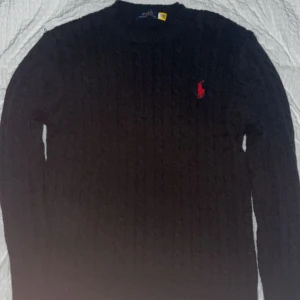 Svart stickad tröja från Polo Ralph Lauren - Kabelstickad svart tröja från Polo Ralph Lauren med klassisk röd logga broderad på bröstet. Tröjan har rund halsringning och långa ärmar, perfekt för lager-på-lager under kyliga dagar.