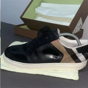 Burberry sneakers med rutigt mönster - Snygga Burberry sneakers med svart läder och mocka framtill, vit sula och klassiskt rutigt tyg i beige, svart och vitt på hälen. Skorna har snörning och en stilren, rund tå. Perfekta för dig som vill ha en lyxig touch på din streetstyle.ganska mycket använda kan sänka pris vid snabb affär 