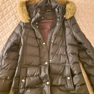 Svart pufferjacka med fuskpäls - Säljer en svart pufferjacka från Marc O'Polo med mysig fuskpäls på huvan. Jackan har quiltad design, dragkedja och knappar framtill samt långa ärmar. Perfekt för kalla dagar och har en snygg, klassisk look.