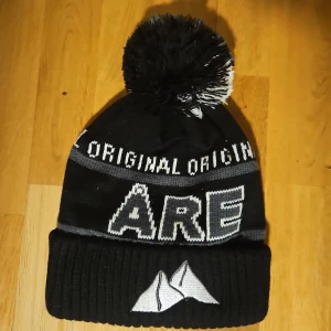Svart Åre mössa med tofs - Snygg svart och grå mössa från Åre Original med stor tofs i svart och vitt. Broderad bergslogga framtill och texten 'ORIGINAL' runt om. Perfekt för kalla dagar och vintersäsongen.