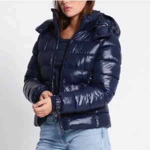 Mörkblå pufferjacka med huva - Snygg mörkblå pufferjacka med glansig finish och huva. Jackan har dragkedja framtill, quiltad design och långa ärmar. Perfekt för kyliga dagar och ger en cool, sportig vibe till din outfit.
