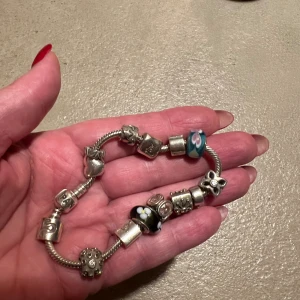 Pandora armband med 12 berlocker och 1 stopp - Unikt Pandora armband i silver med flera berlocker, bland annat hjärtan, stjärnor och blommor. Två glaspärlor sticker ut, en blå med rosa och gula hjärtan och en svart med vita blommor. Perfekt för dig som gillar personliga smycken med detaljer.