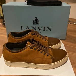 Bruna sneakers från Lanvin i mocka - Snygga bruna sneakers från Lanvin med ovandel i mjuk mocka och tåparti i blankt skinn. Skorna har rund tå, snörning och en beige platt sula som ger en clean look. Perfekta för dig som gillar exklusiva och stilrena sneakers med lyxig känsla.
