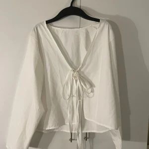 Vit omlottblus med snörning - Säljer en vit blus med omlott-design och djup v-ringning. Blusen har långa ärmar och knyts framtill med snörning, ser inte lika mycket ut med knytningen när man har den på! Liten sminkfläck