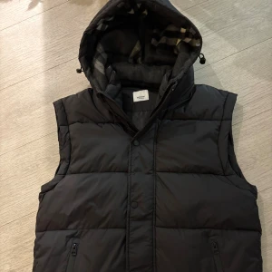 Burberry Jacka - Svart dunväst från Burberry med avtagbar huva och dragkedja framtill. Västen har quiltad design, två fickor med dragkedja och klassisk Burberry-logga på bröstet. Insidan av huvan har ett rutigt mönster i grå toner. Perfekt för lager-på-lager-stil. Storlek M, passar S också men kanske lite för lång för S användare. Mycket Bra skick man kan säga att den är ny. Prislapp finns. 