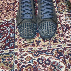 Christian Louboutin svarta sneakers med nitar - Svarta sneakers från Christian Louboutin med klassiska röda sulor och svarta nitar på tån. Skorna har snörning och är tillverkade i mocka, vilket ger en lyxig känsla. Perfekt för dig som vill sticka ut med en edgy och exklusiv stil.