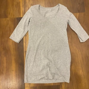 Grå enkel trikåklänning - En basic grå klänning i mjuk trikå med trekvartsärm och rund halsringning. Modellen är rak och kort, perfekt för en avslappnad och stilren look. Passar dig som gillar minimalistiska plagg och vill ha något bekvämt till vardags.